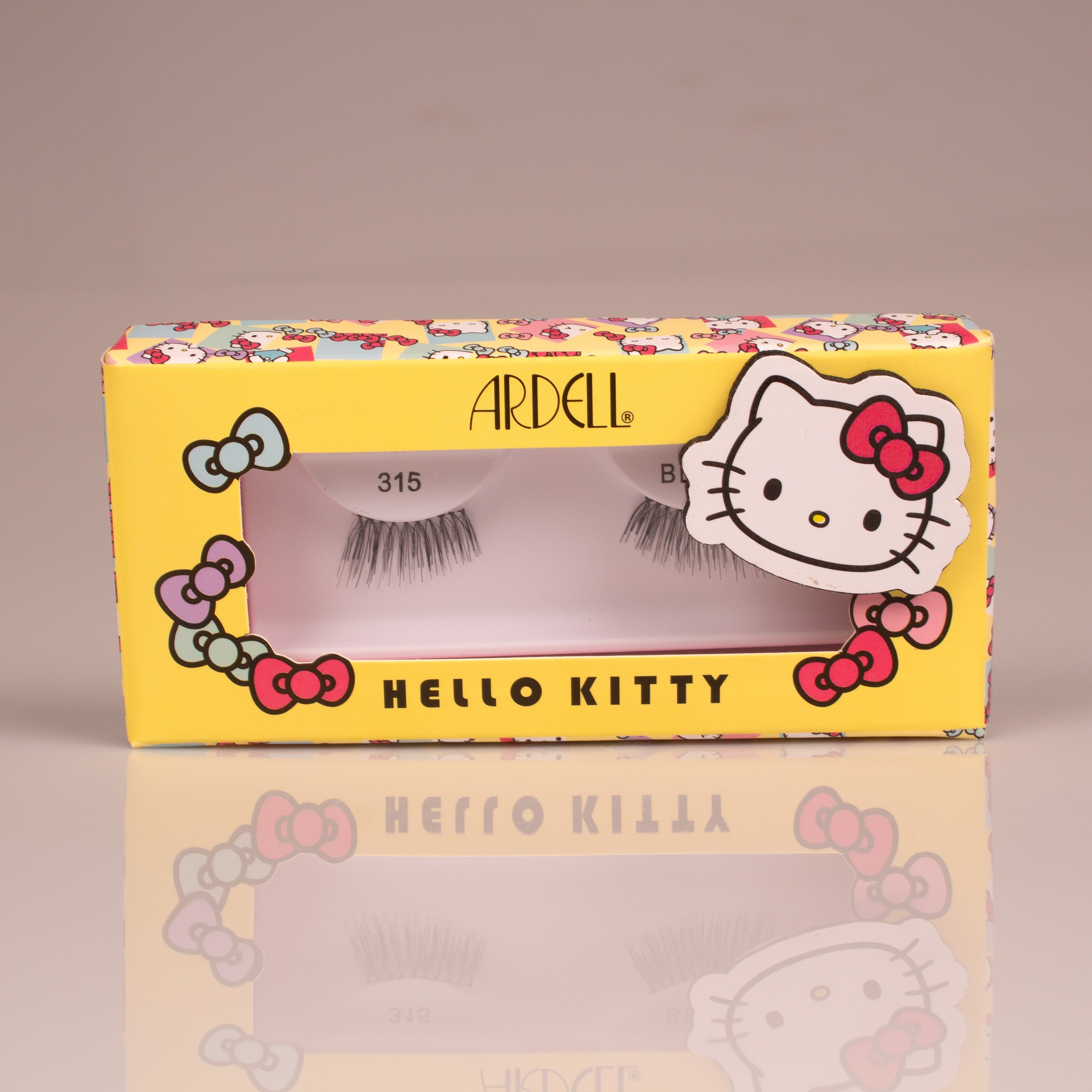 Die ARDELL x Hello Kitty Wimpern im Style 315 stehen vor einem weißen Hintergrund. Sie haben eine gelbe Verpackung, mit dem Hello Kitty Kopf und vielen bunten animierten Schleifen.